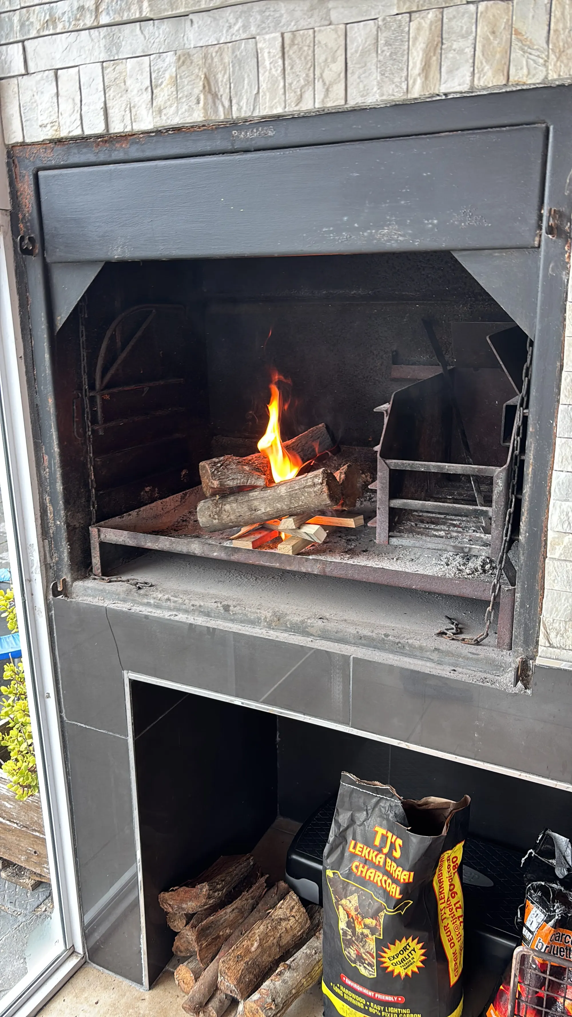 Gemauerte Braai mit brennendem Holzfeuer, darunter das Holzlager mit Scheiten und Holzkohle
