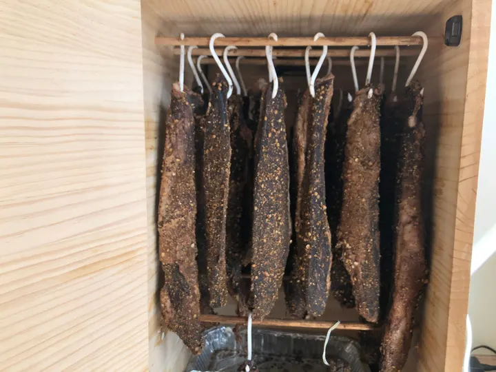 Gewürzte Fleischstreifen hängen an Haken in einem hölzernen Trockenkasten