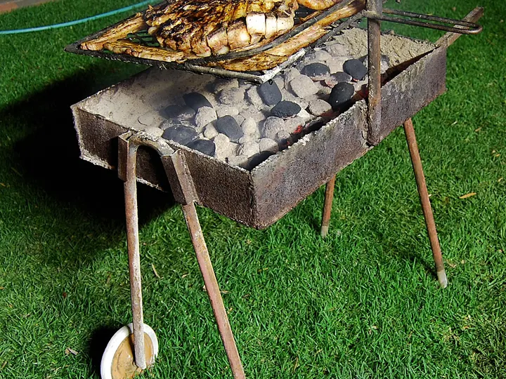 Tragbarer Braai auf vier Beinen mit zwei Rädern, Kohlebett im Blechkasten, Fisch und Wors auf dem Rost