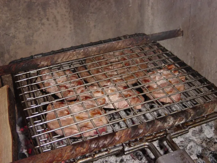 Klapprost mit Fleischstücken zwischen zwei Gittern, am langen Griff über der Glut