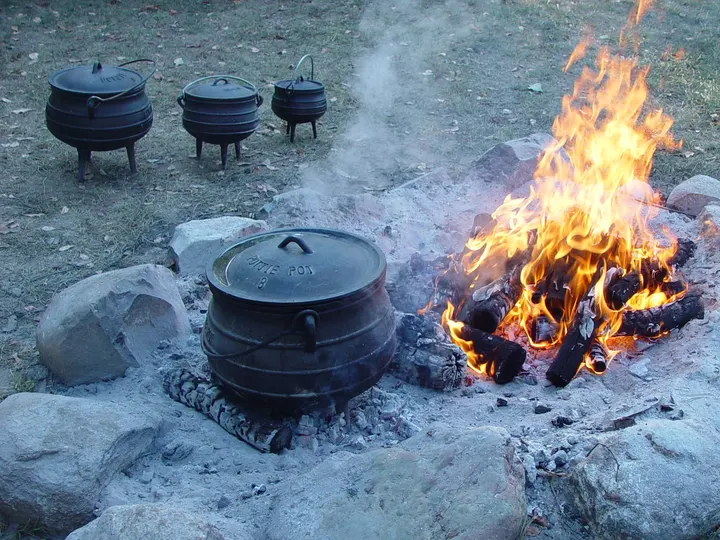 Großer dreibeiniger Gusseisentopf mit Deckel auf der Glut, daneben das Holzfeuer und drei kleinere Töpfe