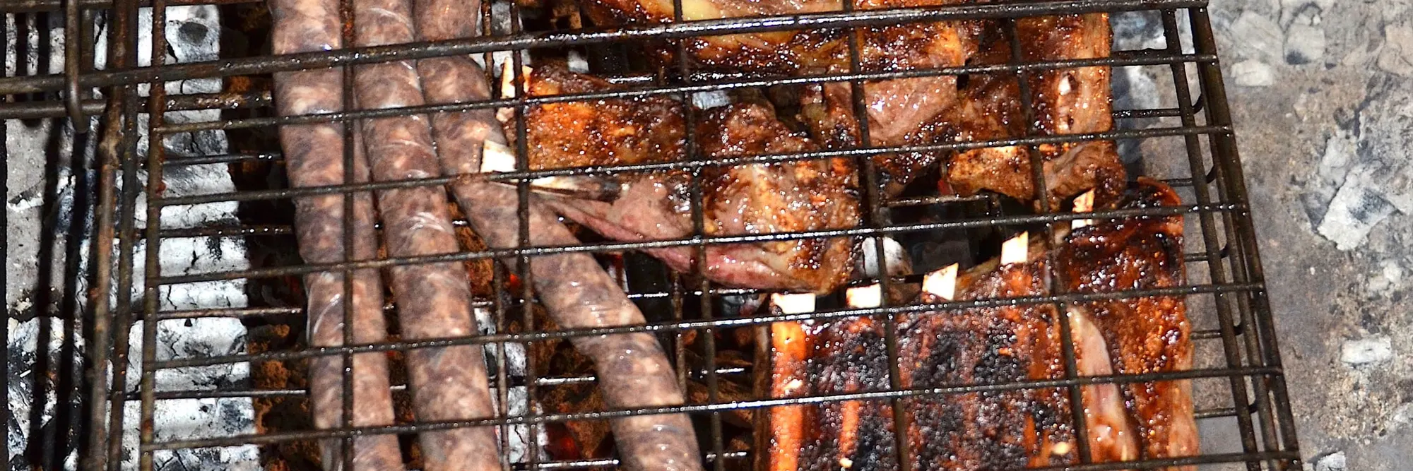 Boerewors in Schlaufen neben Lammrippchen, beides im Klapprost über heller Glut
