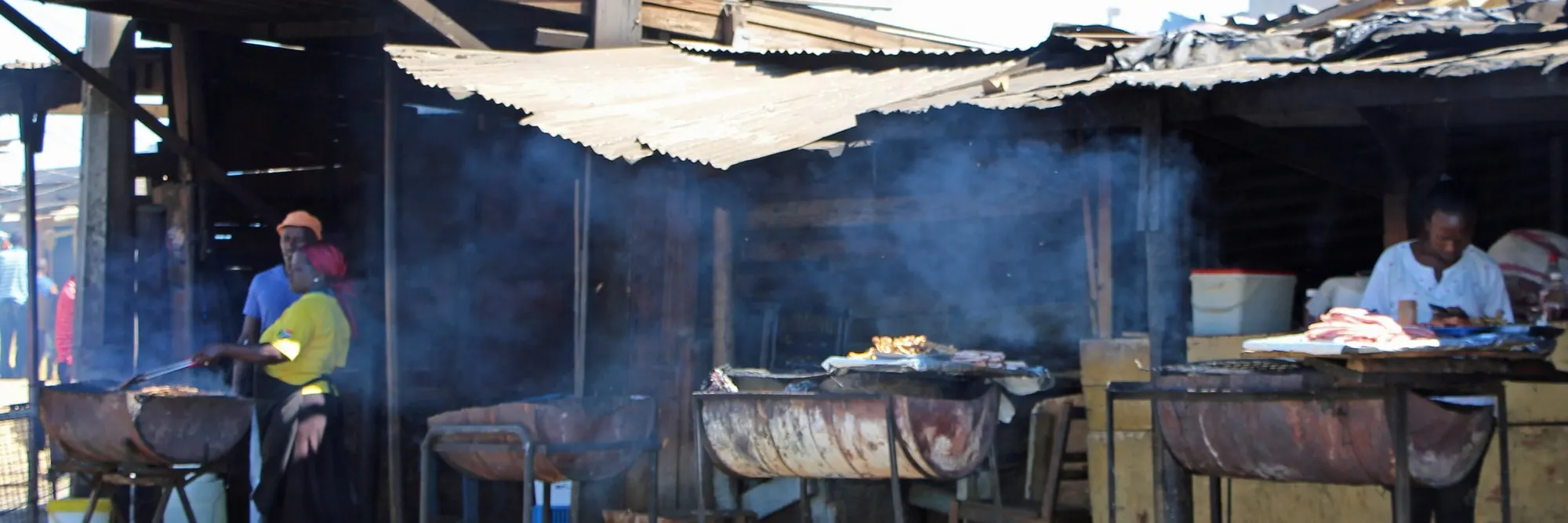 Braai-Stände aus halbierten Ölfässern auf dem Gehweg unter Wellblechdächern, Rauch zieht über die Straße