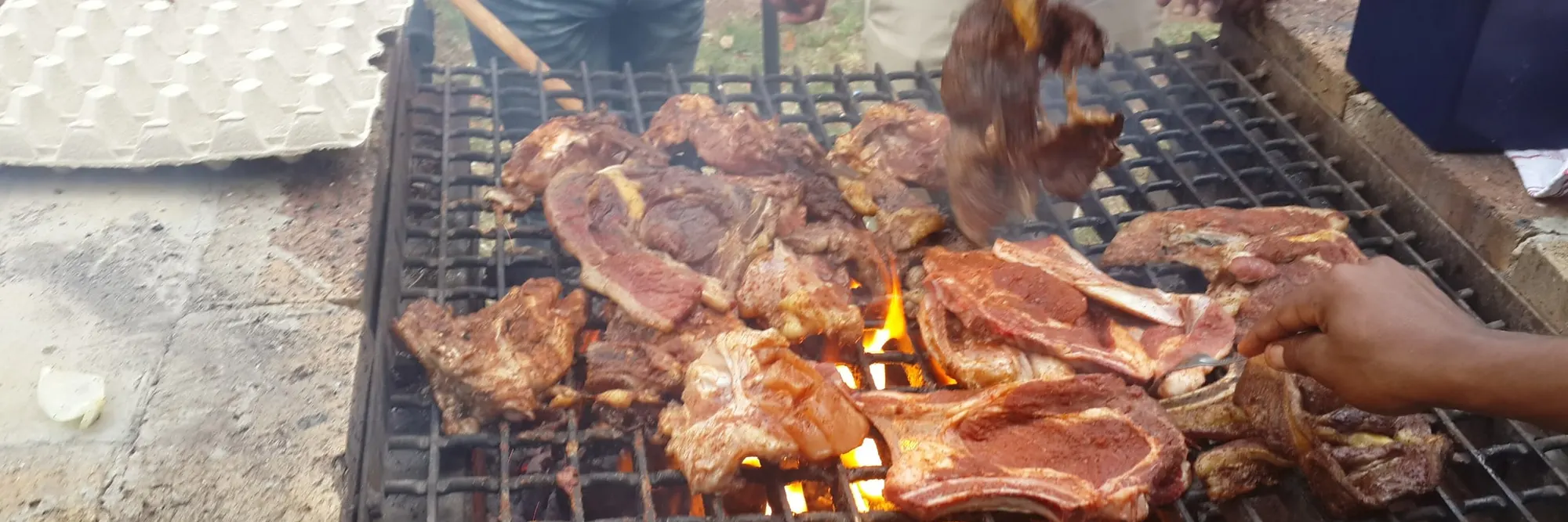 Lammkoteletts auf einem großen Rost, unter dem die Flammen durchschlagen, zwei Paar Hände wenden