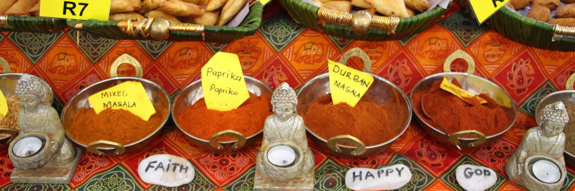 Stahlschalen mit gemahlenem Masala, Paprika und Chili auf gemustertem Tuch, dahinter Körbe mit Samosas